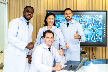 Group of medical practitioners in lab coats gesturing thumbs up while sitting and standing with smiling on camera. Four colleagues discusses the results of an MRI scan of the patient's head
