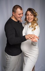 a pregnant woman in white flesh and a man in black clothes on a gray background. Long curly hair, beautiful couple, husband and wife flirting, future parents. stylish pregnancy. portrait
