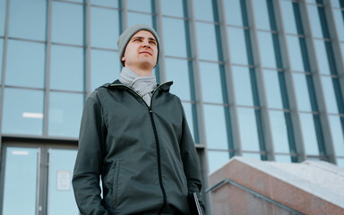 Guy student or freelancer outdoors in business district. Young man in outerwear holding laptop while standing on background of modern building