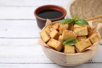 Bowl with delicious fried tofu, basil and sesame seeds on white wooden table, closeup. Space for text