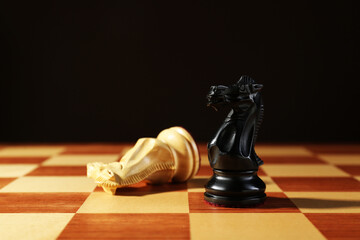 Wooden black knight and fallen one on chessboard against dark background. Competition concept