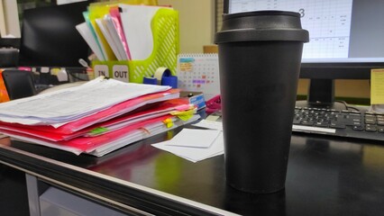 Black tumbler on the black messy desk next to the pile of documents in the office