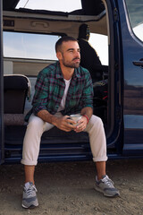 young man sitting in the door of his camper van with a mug in his hand, concept of freedom and nomadic lifestyle