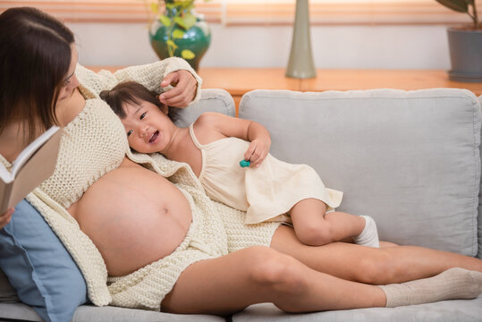 young pregnant mother reading book to her toddler daughter at home,.Image of attractive family pregnant woman and her little daughter smiling and reading book while sitting on sofa at home.