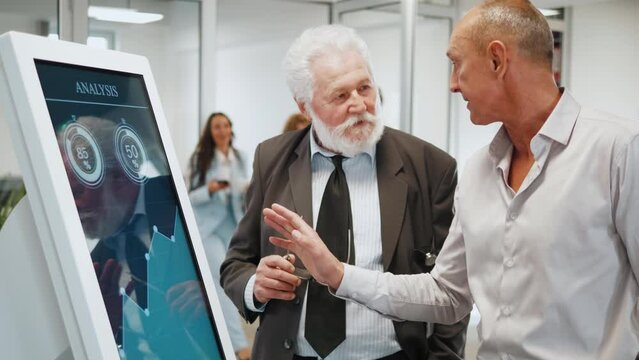 Two old businessmen using a multimedia touchscreen display of interactive kiosk with graph analysis on a screen