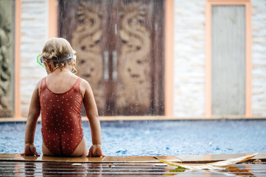 The child sits on the edge of the pool in the rain and is sad in the absence of the sun and good weather.