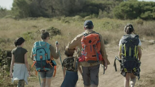 Back view of Caucasian family with backpacks hiking together. Parents and three children walking along path using trekking poles, enjoying vacation together. Handheld shot. Family, travel concept.