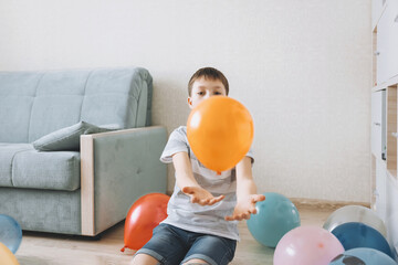 Smiling joyful playful child playing with balloons sitting on floor at come. Boy having fun on kids...