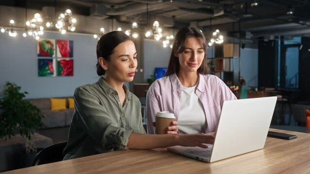Two young beautiful successful women working on laptop sitting in cafe with garland holding cup of coffee smiling talking. Efficient employee and employer manager giving advice. Internet cafe concept.