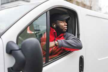 Black young adult delivery guy wearing red pullover and black cap driving white van leaning outside the window to look behind. Horizontal shot. High quality photo