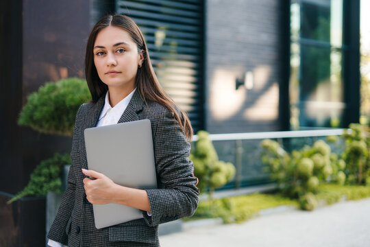 Young brunette business woman standing with laptop in hands, going to office while looking into camera. Working in office, business success, leadership concept