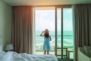 Happy woman wearing blue dress and hat looking outside window to ocean view in morning. Relax,...