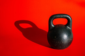 Naklejka na ściany i meble Round black kettlebell with handle on bright red background with shadow. Old heavy metal weight kettlebell for sports, bodybuilding, weightlifting, crossfit. Fitness, power lifting, strength training.