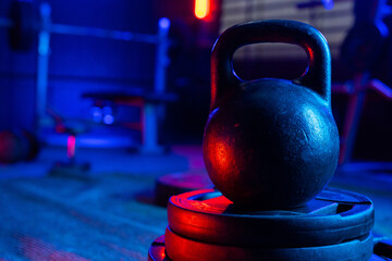 Fototapeta na wymiar Black big kettlebell in the gym with red and blue light. An old round kettlebell with a handle and a barbell rack in the background. Sports equipment for bodybuilding, weightlifting or crossfit.