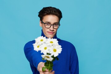 a horizontal portrait of a cute man in black eyeglasses and a blue zip-up sweater, holding out a bouquet of white daisies to the camera with a wide smile on his face