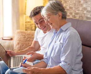 Obraz premium Beautiful young boy sitting on sofa at home helping senior grandmother surf with laptop on the web while she takes notes. Younger generations care for older relatives are teaching computers to use