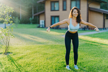 Obraz na płótnie Canvas Slim motivated brunette woman dressed in cropped top and leggings, has workout with dumbbells, poses on green lawn near private house, has perfect body shape. Healthy lifestyle and sport concept