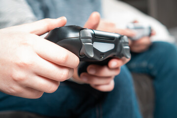 Unrecognizable two teenage boys children sitting at home, holding gaming controller joystick gamepad, playing videogames on game console. Hobby, free time, gaming, entertainment, leisure. Soft focus.