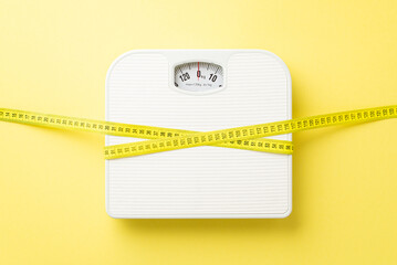 Slimming concept. Top view photo of measuring tape wrapped around scales on isolated pastel yellow...