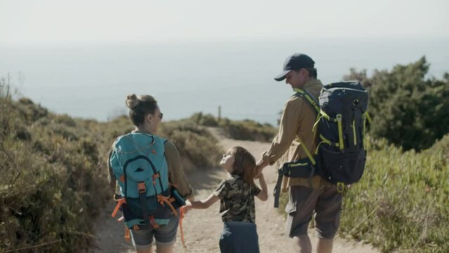 Happy Caucasian family on hiking adventure at seashore. Parents carrying backpacks, holding childs hands, walking along dirt road. Sea background. Back view. Family, leisure activity, travel concept.