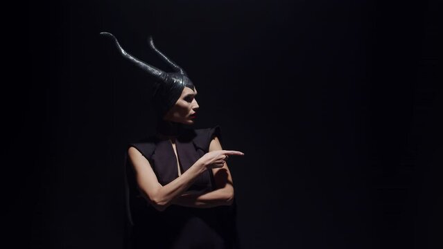 A beautiful evil witch woman with horns stands with her arms crossed over her chest, and then threatens someone with her finger, against a dark background in the studio