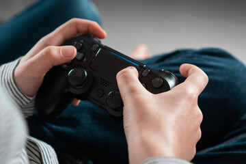 Unrecognizable teenage boy child sitting at home, holding black gaming controller joystick gamepad, playing videogames on console station. Hobby, free time, gaming, entertainment, leisure. Soft focus.