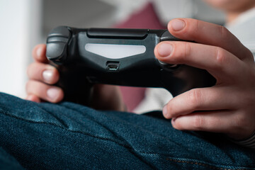 Cropped photo of teenage boy child sitting at home, holding black gaming controller joystick gamepad, playing videogames on console station. Hobby, free time, gaming entertainment, leisure. Back view.