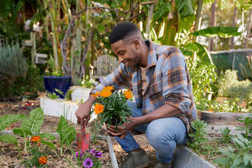 Profile view of happy young black man holding flowers and spade by flowerbed in garden center