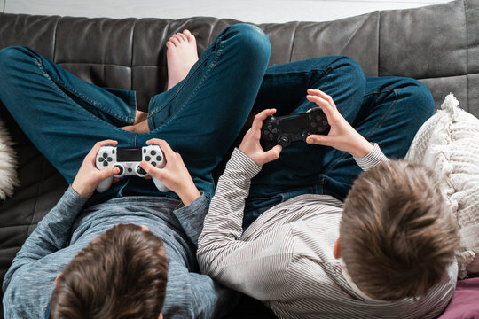 Top view of two teenage boys children sitting on grey sofa at home, holding gaming controller joystick, playing videogames, having fun. Hobby, free time, gaming, entertainment, leisure, friendship.