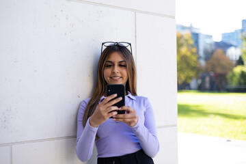 Portrait brown haired woman leans at the wall of the building and reads a message on her smartphone. In the shadow, relaxed position, glasses on top of the head.