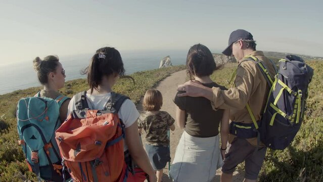 Back view of family backpacking together, hiking on vacation. Parents and children exploring nature while walking along dirt road. Sea background. Fish eye. Slow motion. Hiking, family concept.