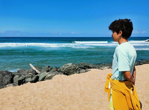 Young Hispanic boy looking at surfers from a seashore