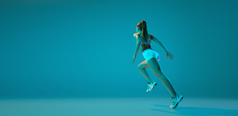 Fototapeta na wymiar Young sportive girl in white sportswear training, running isolated over blue studio background in neon light. Flyer