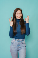 Beautiful caucasian woman with long loose hair in blue sweater and blue jeans posing at studio over turquoise backdrop making victory sign  by both hands. Mockup people in casual.