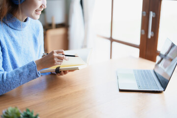 Portrait of a teenage Asian woman using a computer, wearing headphones and using a notebook to study online via video conferencing on a wooden desk at home
