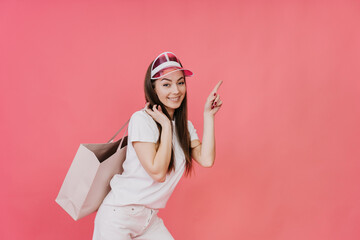 Obraz na płótnie Canvas Playful blonde Italian girl in transparent pink visor, white t-shirt and shorts carrying shopping bag points by index finger at empty space stands against pink studio background. Sale, discount, promo