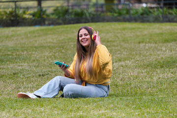 Young girl sitting on the grass with mobile phone headphones listening to music.