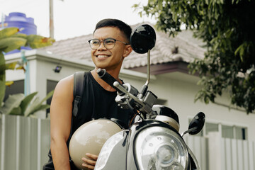 Cheerful Filipino man with eyeglasses sitting on scooter holding vintage helmet. Male motorbike...