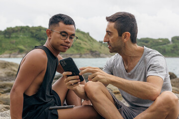 Young man showing cellphone to Filipino friend sitting on rocks by the sea. Filipino and caucasian guy talking about social media holding smart phone. Ethnicity diversity friendship