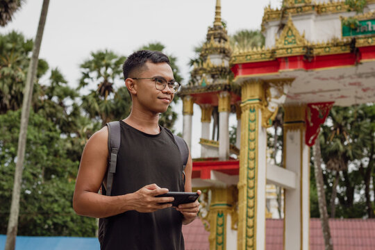 Young Filipino backpacker with mobile phone standing by Buddhist temple in Phuket, Thailand. Male tourist visiting religious building in South East Asia