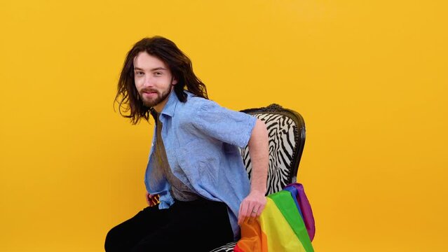 Young caucasian gay man 20s with rainbow striped flag sits in a chair isolated on yellow background. People lifestyle fashion lgbtq concept