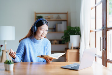 Portrait of a teenage Asian woman using a computer, wearing headphones and using a notebook to study online via video conferencing on a wooden desk at home