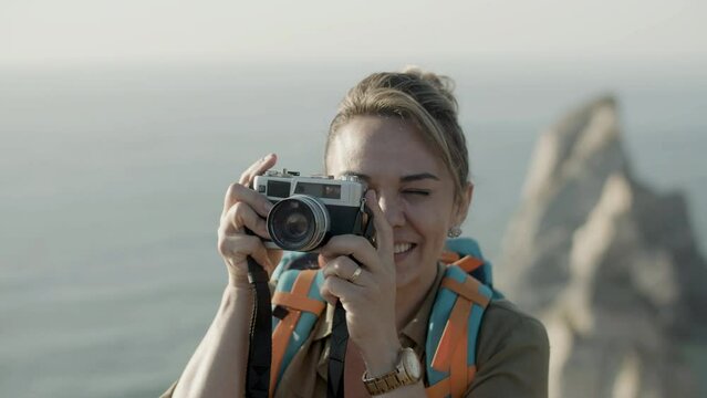 Closeup of beautiful blonde woman taking photo in mountains. Caucasian young female smiling, taking picture with camera against breathtaking sea background. Front view. Photography, travel concept.