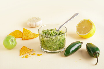 Bowl of tasty green salsa sauce, jalapeno peppers and nachos on color background