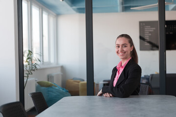 Successful young female leader in a suit with a pink shirt sitting in a modern glass office with a determined smile.