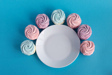 Homemade pink zephyr or marshmallow around white empty plate. Isolated on blue background. Sweet Meringue dessert. Valentineâ€™s or Mothers Day concept. Copy space. Top view