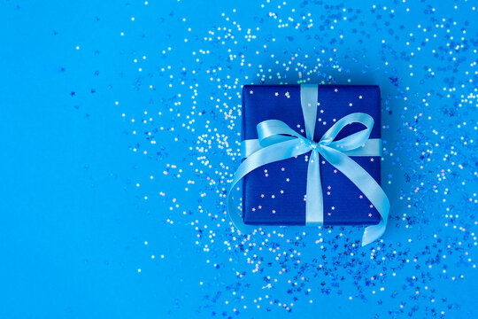 Gift or present box tied with a blue ribbon on blue paper background with sparkle.