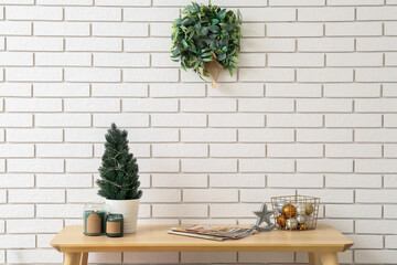 Table with fir tree, magazines, Christmas balls and mistletoe wreath on white brick wall