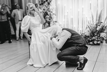 Stylish groom in brown suit pulling out garter poking his head under dress to smiling, laughing,...