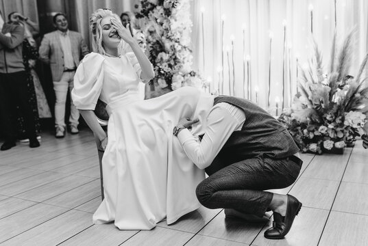 Stylish groom in brown suit pulling out garter poking his head under dress to smiling, laughing, happy blonde bride indoors. Wedding photography, ceremony, Ukraine.